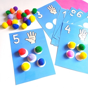 Könnte beinhalten: Ein Set aus blauen Lernkarten zum Zählen mit Zahlen, Handillustrationen mit Fingern und weißen Kreisen zum Platzieren kleiner, bunter Pompons. Die Karten zeigen die Zahlen 3, 4, 5, 6 und 9 mit den entsprechenden Mengen an Pompons.