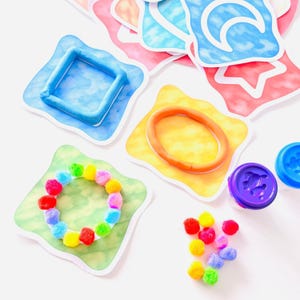 May include: A collection of colourful craft activity cards featuring watercolour-style backgrounds with shapes like a blue square, orange oval, and green circle. One card displays a ring of small multicoloured pom-poms, while others show moon and star outlines.