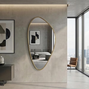 Puede incluir: Un espejo de forma irregular con marco dorado cuelga de una pared gris. El espejo refleja un salón moderno con un sofá gris, una mesa de centro redonda y arte abstracto. Una obra de arte enmarcada está a la izquierda del espejo.