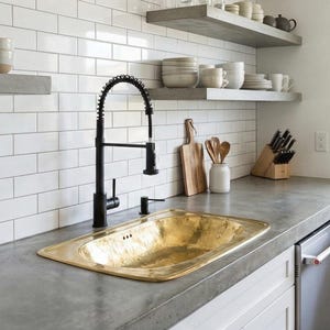 Könnte beinhalten: Eine Küche mit einer goldfarbenen Spüle, einem schwarzen Wasserhahn und weißen U-Bahn-Fliesen. Arbeitsplatten aus Beton und offene Regale zeigen Geschirr und Küchenutensilien. Das Waschbecken ist rechteckig.