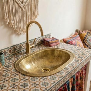 Pode incluir: Uma pia oval de latão martelado com uma torneira correspondente está embutida em uma bancada com azulejos estampados. Uma pilha de toalhas coloridas fica perto da torneira. A pia faz parte de um acessório de banheiro ou cozinha.