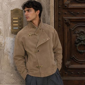May include: A tan suede jacket featuring a high collar, long sleeves, and a front closure secured by three gold-toned metal toggle clasps. The jacket has a structured fit with visible stitching details along the shoulders and chest area.