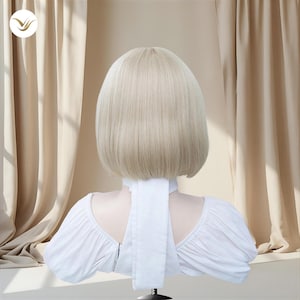 May include: A blonde bob wig with a blunt cut, displayed on a mannequin. The wig is a light, neutral blonde color. The mannequin is wearing a white blouse with a high collar and a long white tie.