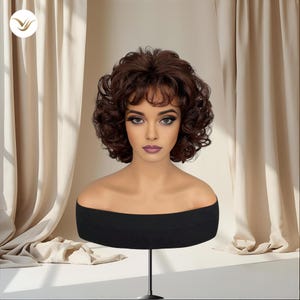 May include: A brown, curly wig with bangs is displayed on a black mannequin head. The wig has a voluminous style and is set against a backdrop of beige curtains. The mannequin is on a black stand.