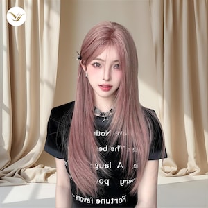 Op de afbeelding: Een persoon met lang, steil, roze haar en een zwart t-shirt met witte tekst. De persoon draagt een zilveren ketting en oorbellen. De achtergrond heeft beige gordijnen.