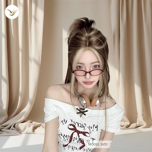 May include: A woman wearing a white off-the-shoulder top with black text and a bow detail. She has brown and blonde hair, red-framed glasses, and headphones around her neck. The background features beige curtains.