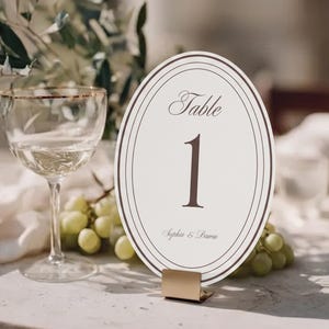 Może przedstawiać: Owalna karta z numerem stołu zawierająca napis Table 1 oraz imiona Sophia & Daren ciemnobrązową kursywą. Karta ma kremowe tło z ciemnobrązowymi koncentrycznymi owalnymi obwódkami i jest umieszczona w złotym metalowym stojaku.
