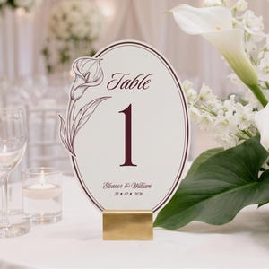 May include: An oval-shaped table number card featuring a dark red calla lily illustration, the word Table, the number 1, and the text Eleanor & William 28 • 12 • 2026. The card is held upright by a rectangular gold-colored metal stand.