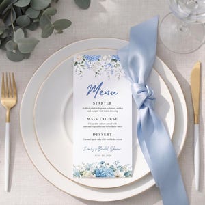 May include: A white menu card with blue floral illustrations at the top and bottom, featuring the text Menu, Starter, Main Course, Dessert, Emily's Bridal Shower, and June 20, 2026, placed on a white plate with gold trim and a light blue silk napkin.