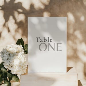 以下が含まれることがあります： 「Table ONE」の文字が黒とグレーで書かれた白いテーブルナンバーカード。カードは明るい木製のブロックの上にあり、左側には白い花と緑の葉があります。木の影が背景に映し出されています。