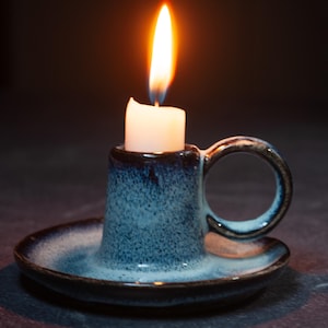 May include: A lit white candle in a blue ceramic candle holder with a handle and a small dish. The candle flame is bright orange and yellow against a dark background. The candle holder has a speckled blue glaze.