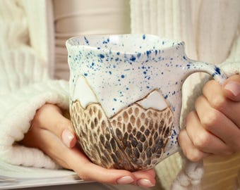 Może przedstawiać: Ręcznie robiony ceramiczny kubek z motywem górskim. Kubek ma białą podstawę z niebieskimi plamkami i teksturowany brązowy łańcuch górski. Uchwyt jest zakrzywiony.