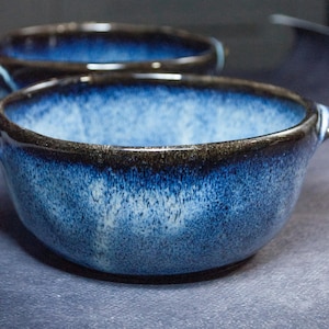 Può includere: Due ciotole in ceramica con una glassa blu intenso e un bordo marrone scuro. Le ciotole hanno una consistenza macchiettata e piccole maniglie. Le ciotole sono probabilmente per servire zuppe o altri piatti.