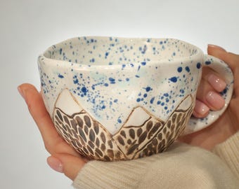 Może przedstawiać: Ręcznie robiony ceramiczny kubek z motywem górskim. Kubek ma białą podstawę z niebieskimi rozpryskami i brązowym wzorem górskim na dole. Ma zaokrąglony uchwyt i jest trzymany w dłoniach osoby.