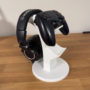 May include: A white stand designed to hold a gaming controller and headphones. A black controller rests on the stand, alongside black over-ear headphones. The stand has a circular base and a curved design. The headphones have the "Audio Technica" logo.