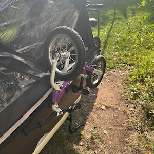 Peut inclure: Remorque de vélo violette et noire avec deux roues. La remorque a un auvent noir et une base blanche et noire. Les roues sont noires avec des rayons argentés. La remorque est sur une zone herbeuse.