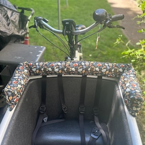 Könnte beinhalten: Ein weißes Lastenrad mit schwarzem Innenraum und einem gemusterten Kissen. Das Kissen zeigt ein Design aus Pilzen, Blättern und anderen Naturelementen. Das Fahrrad hat einen schwarzen Sitz und Sicherheitsgurte.