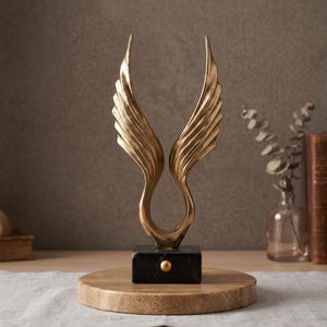 May include: A gold-colored metal sculpture featuring two symmetrical, upward-curving wings attached to a rectangular black marble base with a small gold circular accent, all resting on a circular tan stone pedestal.