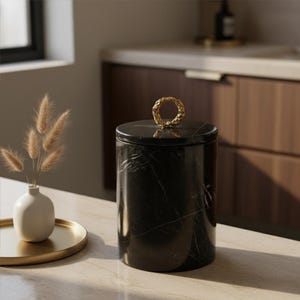 May include: A black marble canister with a gold-colored ring handle. The canister has white veining and sits on a light-colored surface. A small white vase with dried flowers and a gold tray are in the background.