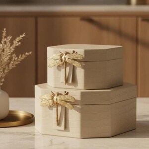 Könnte beinhalten: Zwei beige, sechseckige Schmuckkästchen, die übereinander gestapelt sind. Jede Box hat einen goldenen Libellenakzent. Die Boxen haben ein strukturiertes, krokodilartiges Muster. Die Boxen stehen auf einer Marmoroberfläche.