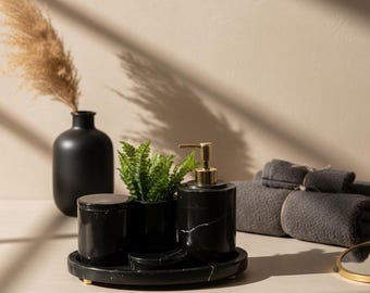 Può includere: Un set da bagno in marmo nero con dettagli dorati, tra cui un dosatore di sapone, un bicchiere e un vassoio, è esposto con una piccola pianta in vaso e asciugamani grigi arrotolati. Un vaso nero con fiori secchi è sullo sfondo.