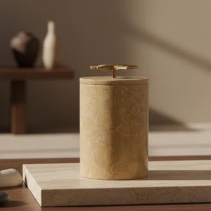 May include: A cylindrical beige marble container with a lid and a gold-coloured floral handle. The container is on a rectangular marble surface. Other decorative items are in the background.