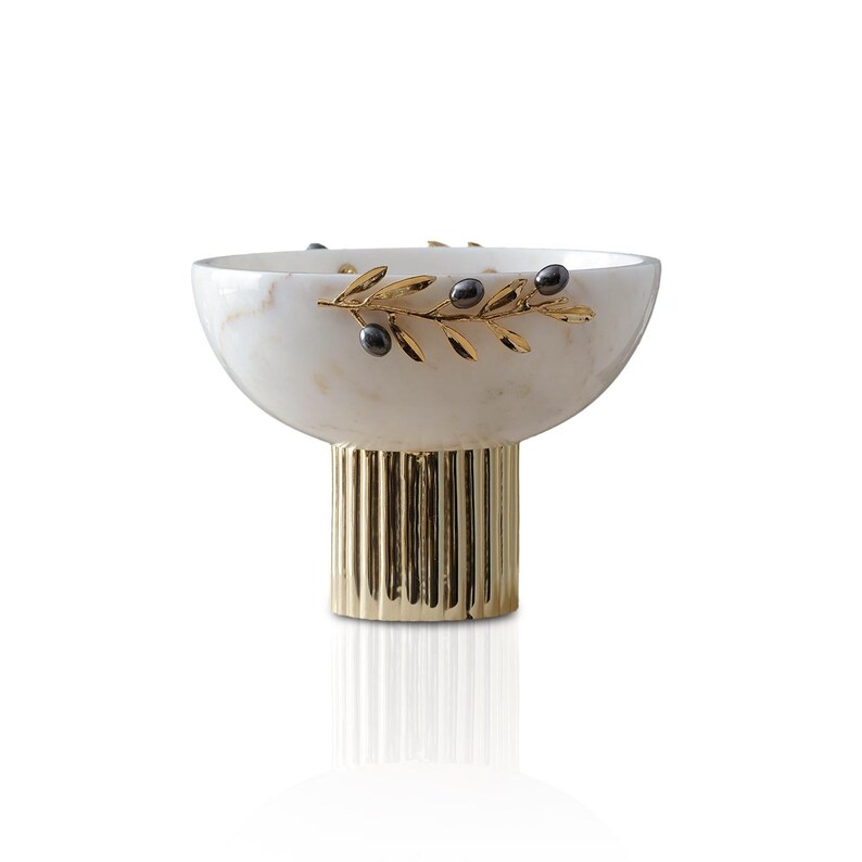 May include: A decorative bowl with a white marble finish, featuring a gold-colored base and olive branch accents. The bowl has a rounded shape and is supported by a fluted, gold-toned pedestal. The olive branch design includes black olive details.