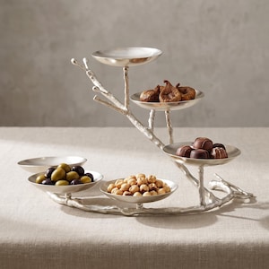 Op de afbeelding: Een zilverkleurige metalen etagère met een takachtige structuur die vijf ondiepe ronde schaaltjes ondersteunt. De schaaltjes bevatten diverse etenswaren, waaronder gedroogde vijgen, diverse chocolaatjes, groene en zwarte olijven en hazelnoten.