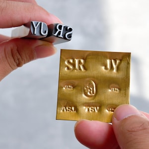 May include: A metal stamp tool with the letters SR and JY engraved on the end, held above a square brass plate. The brass plate features multiple embossed impressions of the letters SR, JY, TSV, and ASL, along with a central oval logo mark.