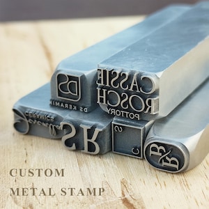 Könnte beinhalten: Eine Sammlung von individuellen Metallstempeln in verschiedenen Formen und Größen mit eingravierten Texten und Logos wie CASSIE ROSCH POTTERY, DS KERAMIK und anderen Monogrammen. Diese silberfarbenen Werkzeuge haben flache, erhabene Oberflächen.