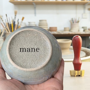 May include: A handmade ceramic bowl with a gray and blue glaze, the word "mane" is stamped on the bottom. A red-handled stamp with a brass head is next to the bowl. The bowl is held in a person's hand.