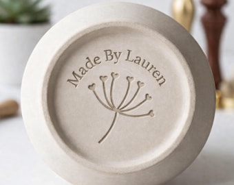 Könnte beinhalten: Nahaufnahme des Bodens einer hellbeigen Keramikschale. Die Worte "Made By Lauren" sind um ein Blumendesign herum eingraviert. Die Schale hat eine abgerundete Form und eine leicht strukturierte Oberfläche.