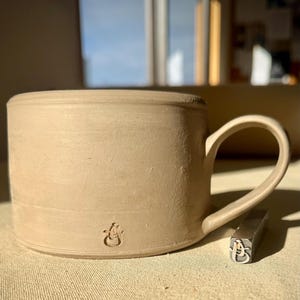 May include: A beige ceramic mug with a smooth, matte finish and a single curved handle. The mug features a small, stamped logo near the base. Beside the mug sits a metal stamping tool displaying the same logo, which consists of the letters J and S.