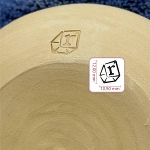 May include: A beige ceramic pottery surface featuring an impressed stamp of a geometric shape containing the letter r. The text MARK YOUR POTTERY is visible at the top. A graphic shows the stamp dimensions of 12.00 mm by 10.90 mm.