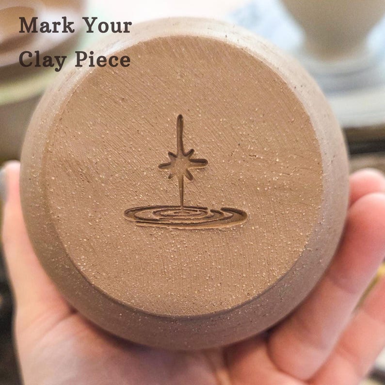 May include: A circular tan clay stamp featuring a debossed starburst icon above concentric ripples. The text 'Mark Your Clay Piece' is printed in the upper left corner. The stamp has a textured, granular surface and a rounded, smooth edge.