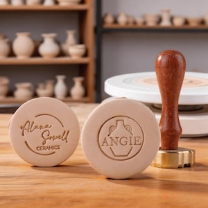 Może przedstawiać: Dwie okrągłe, beżowe pieczątki ceramiczne z wygrawerowanym tekstem i logo. Jedna pieczątka zawiera napis "Alana Sowell CERAMICS" w okrągłym wzorze. Druga pieczątka zawiera imię "ANGIE" wewnątrz konturu wazonu. Widoczna jest również pieczątka z drewnianym uchwytem.