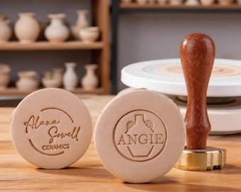 May include: Two round, beige ceramic stamps with engraved text and logos. One stamp reads "Alana Sowell CERAMICS" in a circular design. The other stamp features the name "ANGIE" inside a vase outline. A wooden handled stamp is also visible.