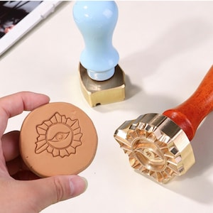 May include: A brass wax seal stamp with a wooden handle featuring a sunburst and eye design, a light blue handled stamp, and a round tan wax impression showing the same sunburst and eye motif, all displayed on a light surface.