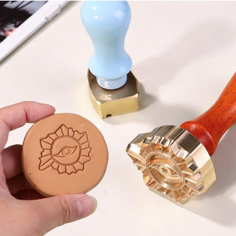 May include: A brass wax seal stamp with a wooden handle featuring a sunburst and eye design, a light blue handled stamp, and a round tan wax impression showing the same sunburst and eye motif, all displayed on a light surface.