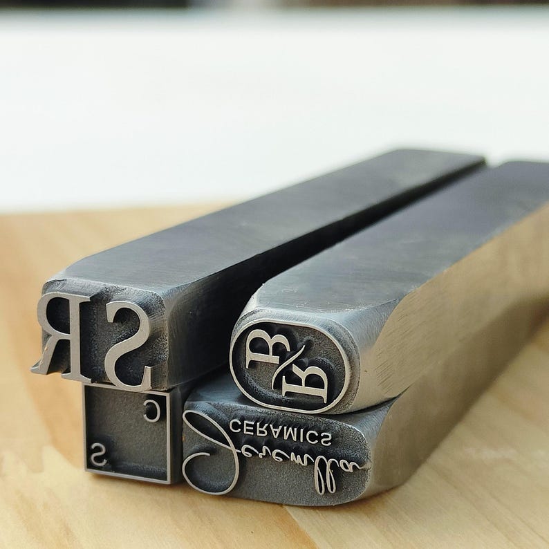 May include: Close-up of several metal stamps with various designs. One stamp features the letters "RB" inside an oval, another has the word "Ceramics" in reverse, and others have numbers and letters. The stamps are on a wooden surface.