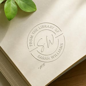 Peut inclure: Un livre crème avec un emblème circulaire en relief indiquant "FROM THE LIBRARY OF SARAH WILLIAMS" avec les initiales "SW" au centre. Le numéro "108" est en dessous du texte. Une feuille verte est dans le coin supérieur gauche.