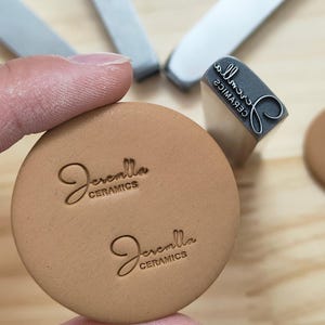 May include: A close-up of a tan ceramic stamp with the name "Jeremlla Ceramics" in a cursive font. The stamp is pressed into a round, tan ceramic piece. A gray stamp and other tools are in the background.