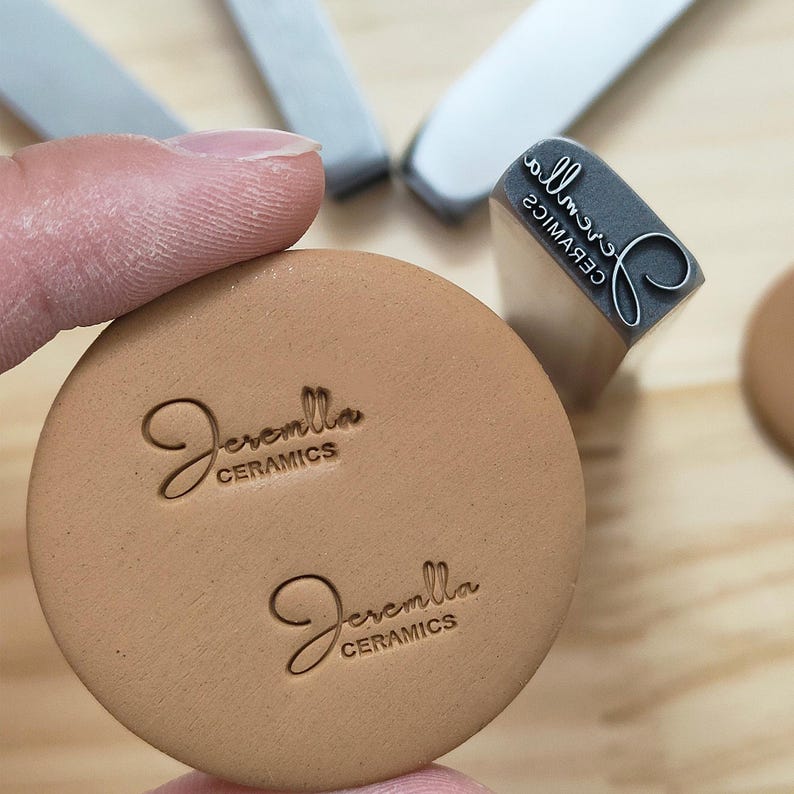 May include: A close-up of a tan ceramic stamp with the name "Jeremlla Ceramics" in a cursive font. The stamp is pressed into a round, tan ceramic piece. A gray stamp and other tools are in the background.