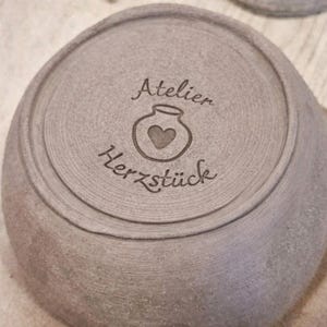 Op de afbeelding: Een grijze keramische kom met de woorden "Atelier Herzstück" en een hartvormig ontwerp geëtst in de bodem. De kom heeft een afgeronde vorm met concentrische ringen bij de rand.
