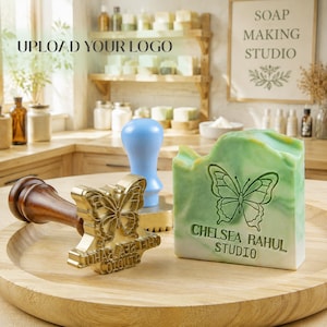 May include: A brass soap stamp with a dark wooden handle featuring a butterfly design and the text CHELSEA RAHUL STUDIO, positioned next to a rectangular bar of green and white marbled soap embossed with the same butterfly and text logo.