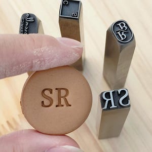 Puede incluir: Un círculo de cuero marrón claro estampado con las letras "SR" es sostenido por los dedos de una persona. En el fondo hay cuatro sellos de letras metálicas, incluyendo sellos con las letras "B", "C" y "2".