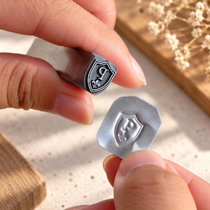 Puede incluir: Una herramienta de estampado de metal plateado con un diseño de escudo que incluye la letra P y un símbolo de flor de lis. La herramienta se muestra junto a una pequeña pieza de metal cuadrada que ha sido marcada con el mismo diseño de escudo, letra P y flor de lis.