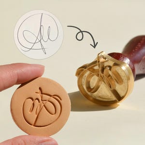 Puede incluir: Un sello de latón con un diseño de firma personalizado junto a una impresión estampada en un círculo beige. El sello se muestra junto a una firma de muestra y una impresión estampada. El sello tiene un mango de madera.