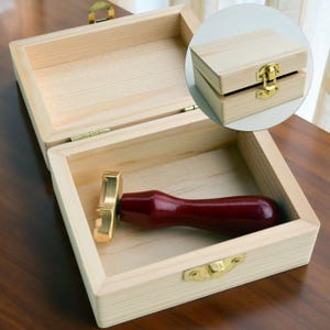 May include: A rectangular light-colored wooden box with a gold-toned metal latch and hinges. Inside the open box lies a wax seal stamp featuring a dark red wooden handle and a brass head. An inset shows the same wooden box in a closed position.