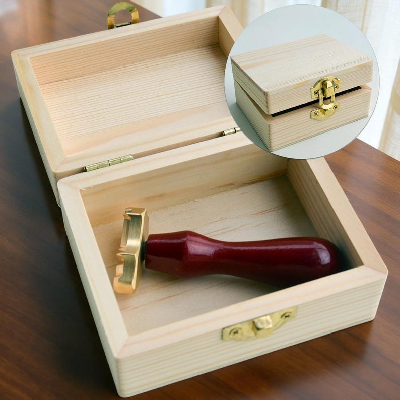 May include: A rectangular light-colored wooden box with a gold-toned metal latch and hinges. Inside the open box lies a wax seal stamp featuring a dark red wooden handle and a brass head. An inset shows the same wooden box in a closed position.